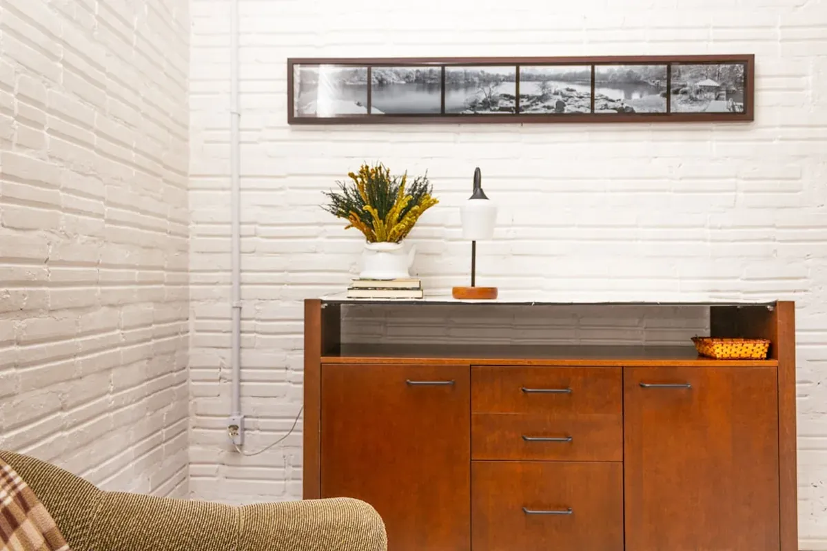 A wooden sideboard cabinet with closed doors and drawers against a white painted brick wall, a lamp and plant on top